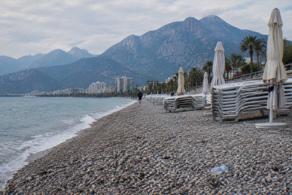 Rows of sunbeds and umbrellas along a pebble shore with a promenade behind them
